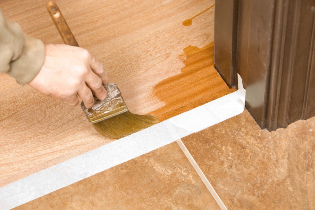 Applying clear polyurethane finish to hardwood flooring with a brush near a tile transition